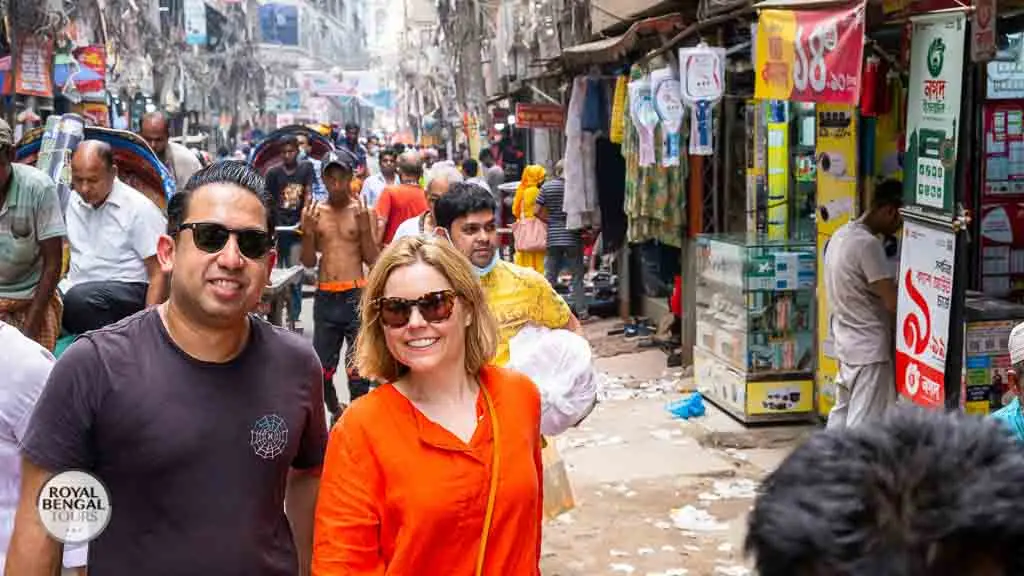 Western couple walking on a busy street lined with shops and colorful signs in Old Dhaka, Bangladesh.