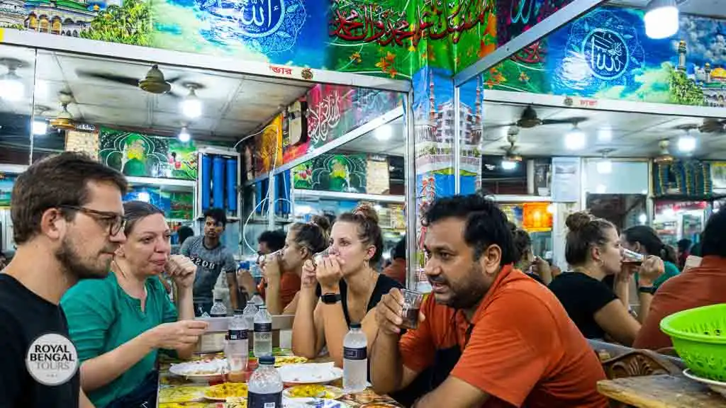 Group at a table with Bangladeshi food at a Dhaka bazaar. Streetside market with vendors and customers.